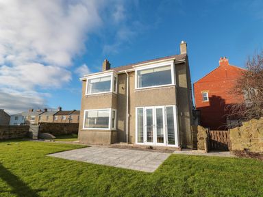 A two-story house with large windows and a patio in front and a lawn at Fairwinds in Newbiggin-By-The-Sea