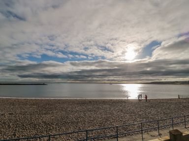 A sandy beach with a few people and dogs near the water with a sun partially covered by clouds at Fairwinds in Newbiggin-By-The-Sea