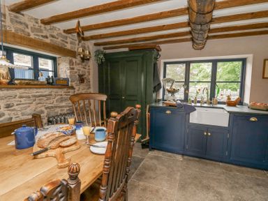 A kitchen with wooden chairs a wooden table with bread and juice stone walls blue cabinets and a window at Ty Sinsir in Tretower