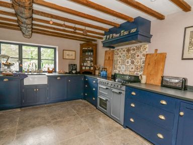 A kitchen with blue cabinets a window above a white sink a stove with patterned tile backsplash and wooden ceiling beams at Ty Sinsir in Tretower