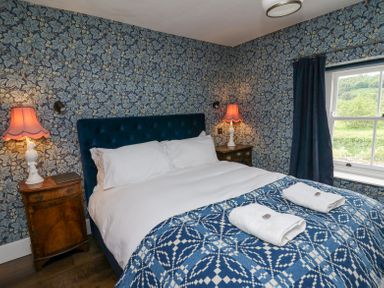 A bedroom with a double bed with white linens and blue patterned blanket with towels on top two bedside tables with lamps and floral wallpaper at Ty Sinsir in Tretower
