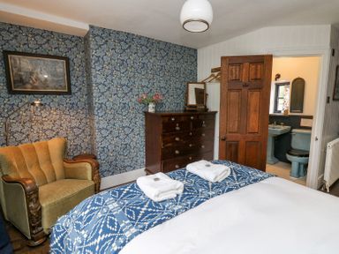 A bedroom with a bed, a blue patterned bedspread, an armchair, a wooden chest of drawers, floral wallpaper, and an open door to a bathroom at Ty Sinsir in Tretower