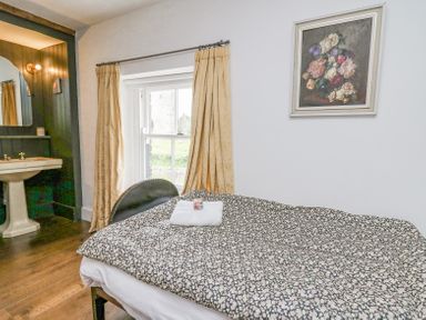 A bedroom with a bed covered in patterned bedding a window with curtains a sink on wooden floor and a floral painting on the wall at Ty Sinsir in Tretower