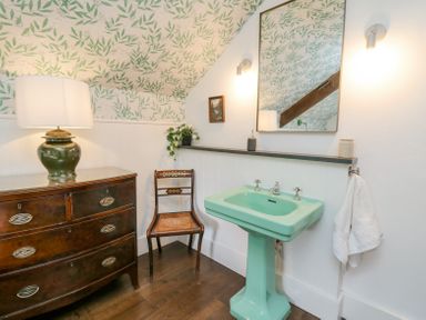 A bathroom with a green sink a wooden chair a dark wooden dresser with a lamp and floral wallpaper at Ty Sinsir in Tretower