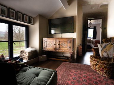 A living room with a television on a wooden cabinet next to a window with folded blankets and an armchair with a patterned pillow at Ty Sinsir in Tretower