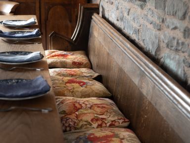 A dining table with plates forks and napkins next to wooden bench seating with patterned cushions against a stone wall at Ty Sinsir in Tretower