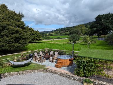 An outdoor area with a hot tub wooden chairs a hammock and a green roofed shed in a grassy field at Ty Sinsir in Tretower