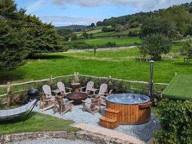 An outdoor seating area with wooden chairs around a fire pit and a hot tub near a green field at Ty Sinsir in Tretower