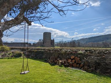 A yard with a swing hanging from a tree a pile of logs a stone wall and ruins in the background at Ty Sinsir in Tretower