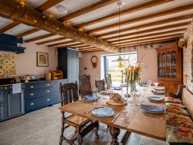 A dining area with a wooden table set with plates glassware and candles and blue kitchen cabinets in the background at Ty Sinsir in Tretower