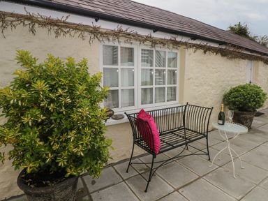 A patio with a black metal bench with pink cushions a small white table with a bottle and glass and two potted plants at Primrose Cottage in Manorbier