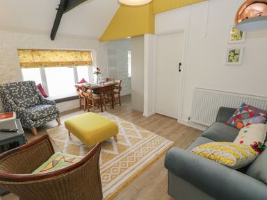A sitting area with a gray sofa, patterned armchairs, a yellow ottoman, a wooden dining table near a window with yellow curtains at Primrose Cottage in Manorbier