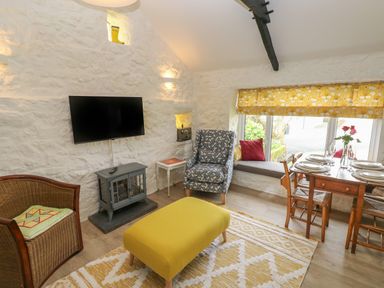 A living room with a yellow ottoman a patterned armchair a wicker chair a dining table set with plates and glasses and a window seat at Primrose Cottage in Manorbier