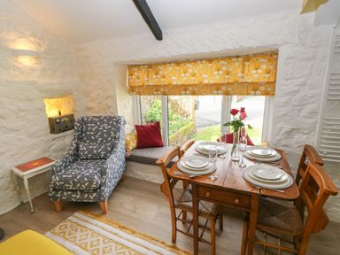 A dining area with a wooden table set with plates and glasses next to a window with yellow floral curtains and a patterned armchair at Primrose Cottage in Manorbier