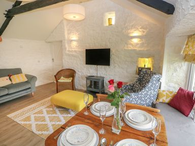 A living room with a dining table set with plates and glasses a yellow ottoman a sofa armchair and a wall mounted TV at Primrose Cottage in Manorbier