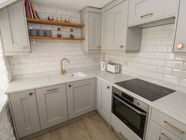 A kitchen with grey cabinets white countertops a sink with brass faucet a toaster kettle stove oven and wooden shelves with mugs and containers at Primrose Cottage in Manorbier