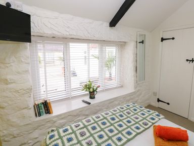 A bedroom with a bed covered by a crocheted blanket an orange towel on the bed a window with blinds a small mirror mounted on the wall books on the windowsill and a mounted television at Primrose Cottage in Manorbier