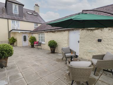 An outdoor patio area with stone flooring, potted plants, wicker chairs with cushions, a large green umbrella and a small table with a bottle and glasses at Primrose Cottage in Manorbier