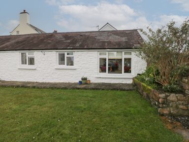 A white stone cottage with three windows a grassy lawn and potted plants at Primrose Cottage in Manorbier