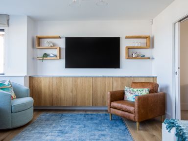 A living room with a mounted flat screen tv wood cabinet shelving and armchairs at 12 Tintagel Terrace in Port Isaac