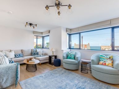 A living room with a beige sectional sofa and blue armchairs near windows showing a sea view at 12 Tintagel Terrace in Port Isaac