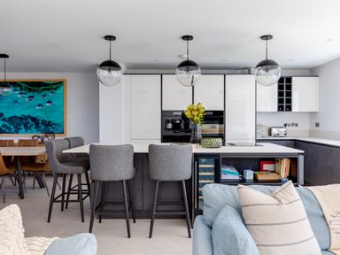 A modern kitchen with a white island and gray stools three pendant lights and a dining table with brown chairs at 12 Tintagel Terrace in Port Isaac