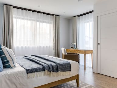 A bedroom with a bed covered in a blue blanket and pillows and a wooden desk with a chair near two large windows at 12 Tintagel Terrace in Port Isaac