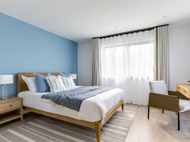 A bedroom with a bed featuring blue and white pillows a wooden nightstand with a lamp a striped rug a window with white curtains and a wooden desk with a chair at 12 Tintagel Terrace in Port Isaac