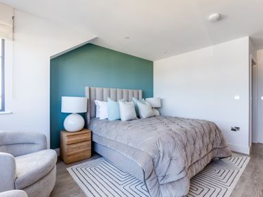 A bedroom with a grey upholstered bed with multiple pillows a wooden bedside table with a lamp a grey armchair and a patterned rug at 12 Tintagel Terrace in Port Isaac