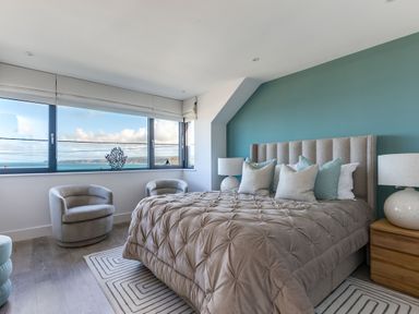 A bedroom with a large bed with pillows two armchairs bedside tables with lamps and a window with a sea view at 12 Tintagel Terrace in Port Isaac