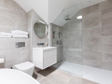 A bathroom with a wall-mounted sink, circular mirror, toilet, folded towels, and a walk-in shower at 12 Tintagel Terrace in Port Isaac