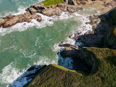 A rocky coastline with waves crashing against cliffs and a small sandy beach at 12 Tintagel Terrace in Port Isaac