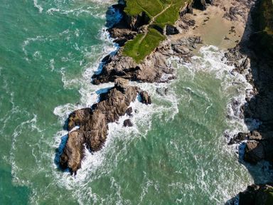 An aerial view of a rocky coastline with green grass and waves crashing at 12 Tintagel Terrace in Port Isaac