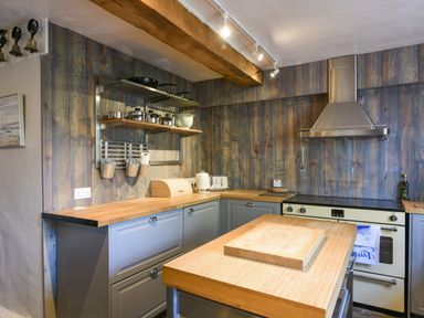 A kitchen with wooden countertops a stove with a hood a wooden chopping board a metal shelf with kitchen utensils and blue cabinets at Braeside Lyme Regis