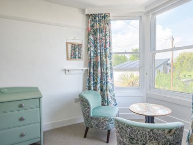 A small seating area with two chairs a round table beside windows with floral curtains and a green chest of drawers at Braeside Lyme Regis