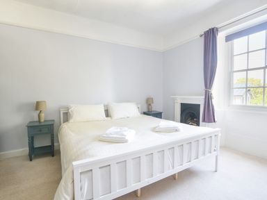 A bedroom with a white bed frame, two bedside tables with lamps, a fireplace, and a window with curtains at Braeside in Lyme Regis