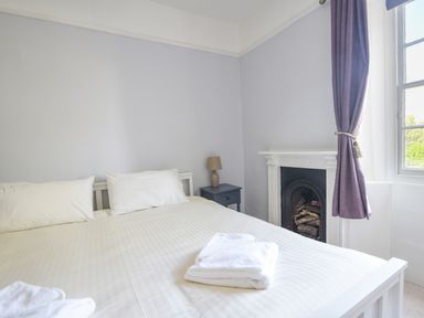 A bedroom with a bed, two pillows, a bedside table with a lamp, a fireplace, and a window with a curtain at Braeside in Lyme Regis
