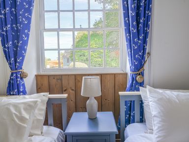 A bedroom window with blue anchor curtains between two single beds and a bedside table with a lamp at Braeside in Lyme Regis