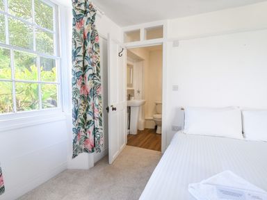 A bedroom with a bed white sheets a window with floral curtains and an open door leading to a bathroom at Braeside in Lyme Regis