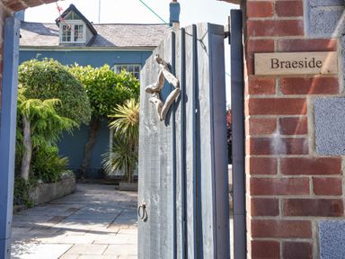 An open wooden gate with a decorative handle leading to a paved courtyard with trees and a blue house in the background at Braeside Lyme Regis