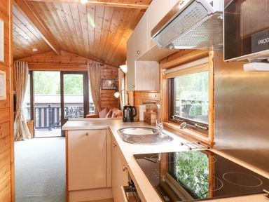 A kitchen area with a sink stovetop and kettle next to a living area with sofa and sliding glass doors at Ennerdale Lodge Burnside Park near Keswick