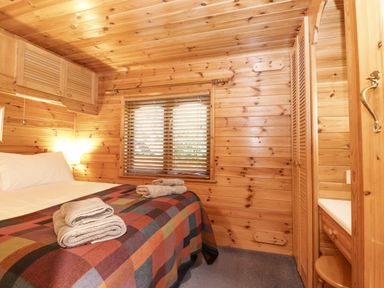 A bedroom with a bed covered by a multicolored checkered blanket with folded towels on top wooden walls a window with blinds and built in wooden cabinets at Ennerdale Lodge Burnside Park near Keswick