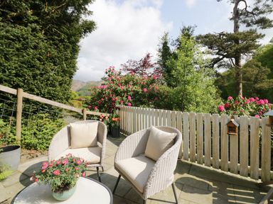 A patio with two wicker chairs a white table with flowers and a wooden fence at Old Farm Cottage in Skelwith Fold