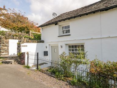 A white cottage with a grey door and small windows surrounded by a low metal fence and stone walls at Old Farm Cottage in Skelwith Fold