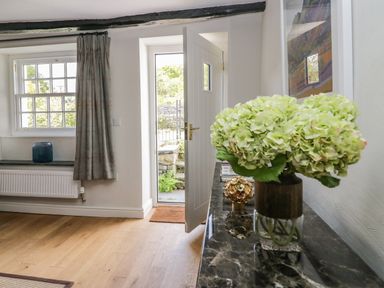 An entryway with an open white door showing a stone wall outside and a table with a vase of flowers and decor at Old Farm Cottage in Skelwith Fold