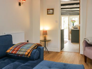 A living room with a blue sofa a checkered blanket a side table with a lamp and an open doorway to a kitchen with flowers at Old Farm Cottage Skelwith Fold