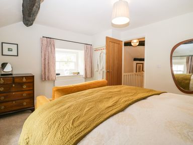 A bedroom with a bed covered with a yellow blanket a wooden dresser a window with pink curtains an open wooden door and a wall mirror at Old Farm Cottage Skelwith Fold