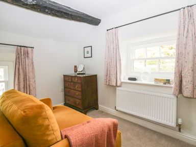 A sitting room with an orange sofa a wooden chest of drawers and two windows with pink curtains at Old Farm Cottage in Skelwith Fold