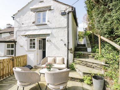 An outdoor patio with wicker chairs and a small table in front of a white house with stone steps and greenery at Old Farm Cottage in Skelwith Fold