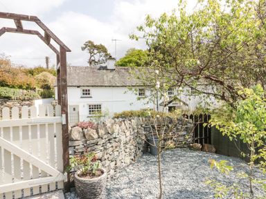 A garden area with small trees gravel ground and stone wall with a white gate at Old Farm Cottage in Skelwith Fold
